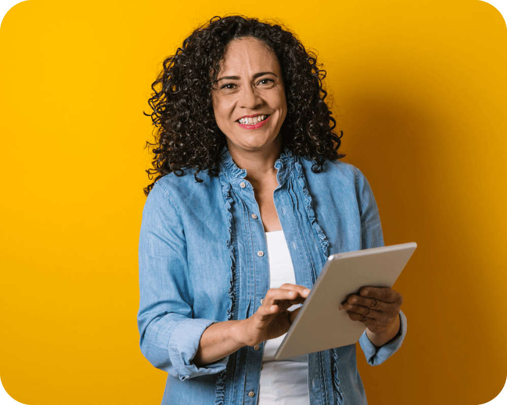 Picture of a lady holding a clip board. She has a white T-shirt and a blue blouse on. She is pictured on a yellow background and is smiling at the camera